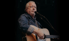 Russell Moore strums his brown guitar in front of a black backdrop.
