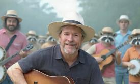 Songwriter Kevin Edwards with his guitar, outside, backed by banjo playing racoons and others