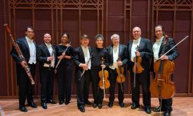 Eight musicians in a winds and strings ensemble, standing in a room with majestic wood paneling