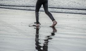 A surfer in a wetsuit walking on shallow water on the shoreline.