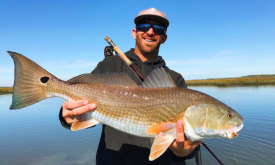 A man in a hat and sunglasses holds a large red drum fish on the waterways of St. Augustine.
