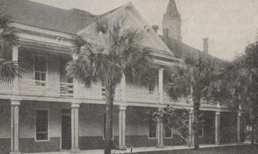 A black and white photograph of the Governor's House in St. Augustine, Florida. It is a three-story building with a wrap-around balcony on the second floor. The bell tower of the Cathedral can be seen in the background above the roof of the Governor's House, showing that the Cameraman was facing north.