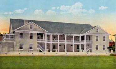 A color-tinted photograph (taken in the 1920s) of the St. Francis Barracks in St. Augustine, Florida. It is a building with three stories. The center section is recessed into a patio on the ground level and a white-railed balcony on the second level. Its walls are beige and its roof is grey.