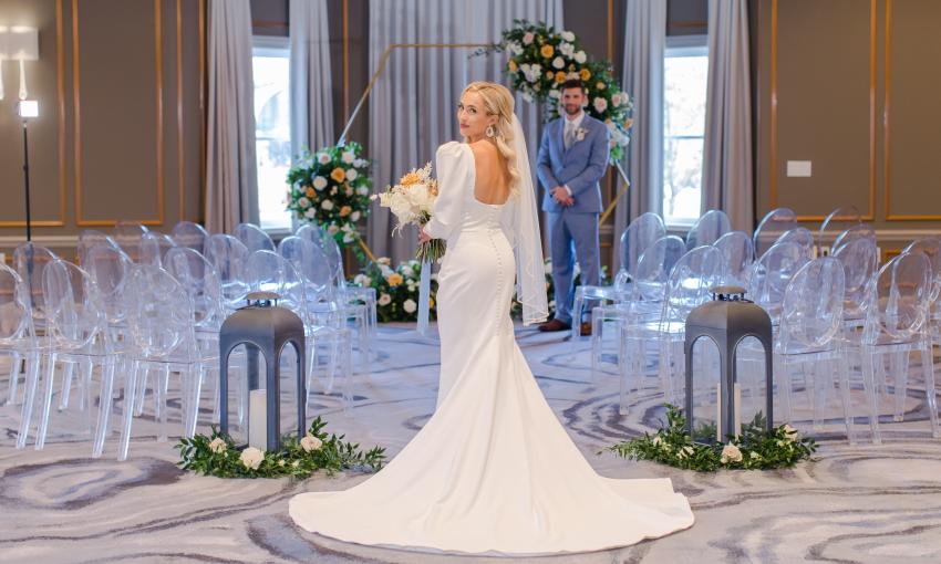 A bride, looking over her shoulder as the groom waits in a hotel room set up for a wedding