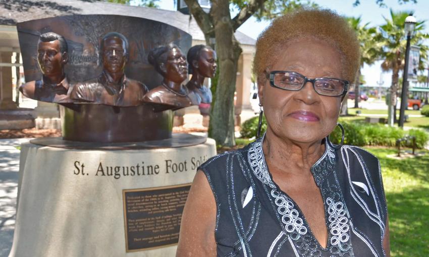 Barbara Vickers (an older Black woman wearing glasses) stands in front of the bronze monument to the St. Augustine Foot Soldiers.