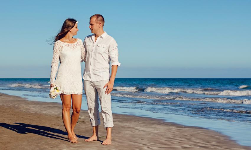 A bride and groom standing on the beach at their wedding, both are barefoot, she is in a short white dress, and he is in a white shirt and rolled-up pants