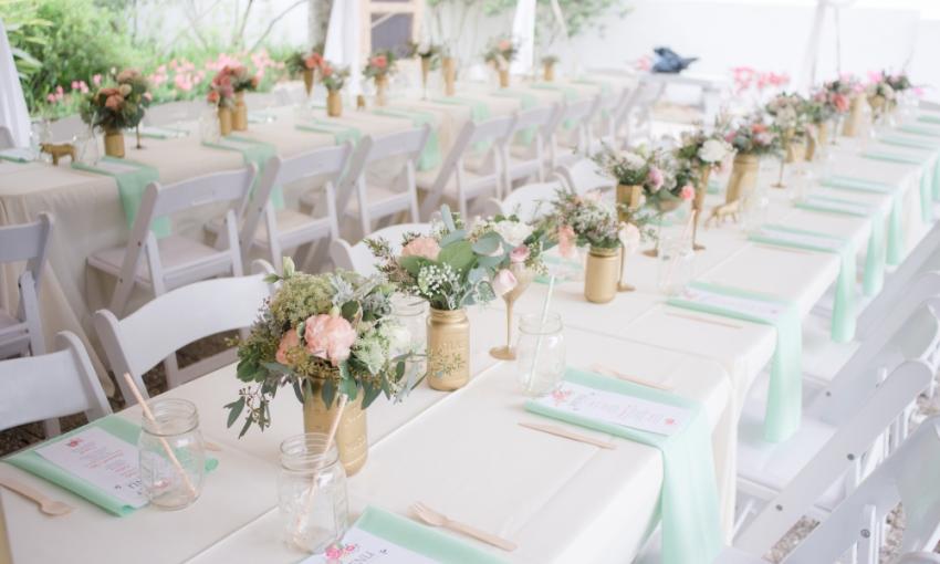 Tables arranged for a wedding reception, with white cloths, aqua napkins, and pink flowers in vases full of greenery