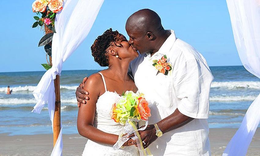 This bride and groom share their kiss under a canopy at the beach, with sand, water, and blue sky in the background