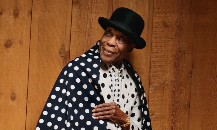 Buddy Guy stands for the camera in a black hat and a polka-dot jacket.